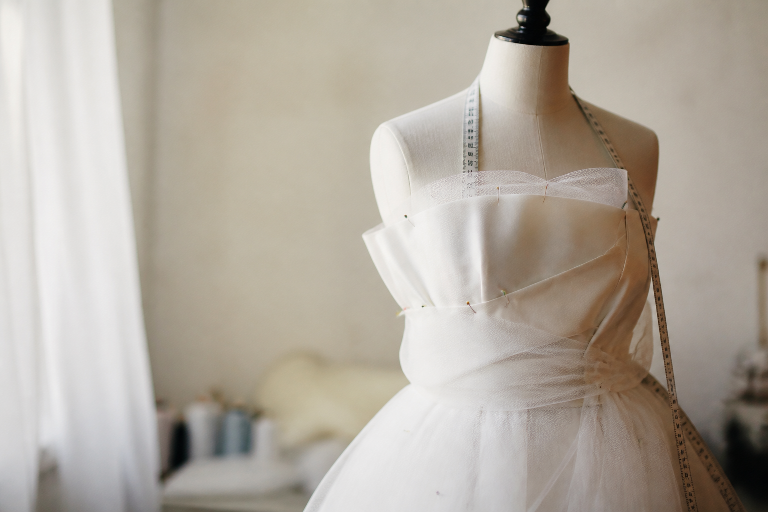 Robe de cérémonie claire suspendue à un mannequin de couture, tulle et satin retenus par des épingles, mètre ruban autour du cou du mannequin